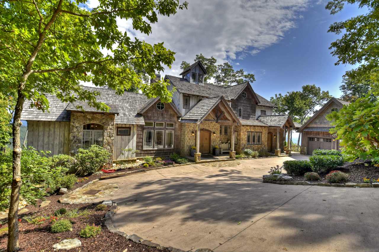 A large stone house with a driveway and a big tree in front of it. 