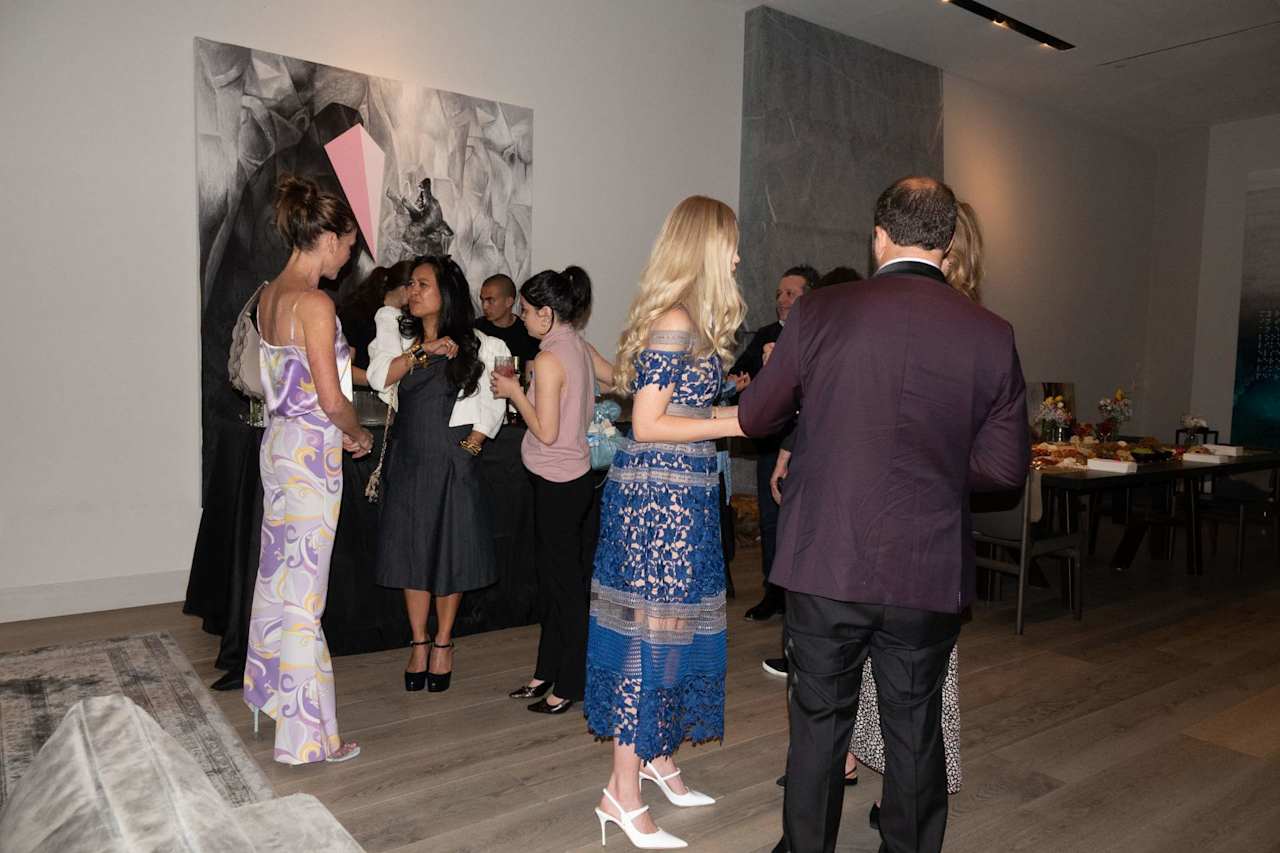 Guests mingle at an indoor event beside contemporary artwork and a refreshment table.