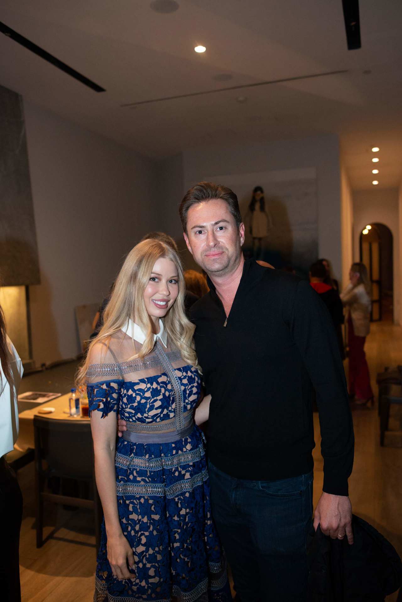 A woman in a blue patterned dress and a man in black pose together at an indoor event.