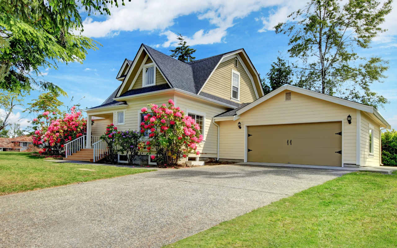 Light yellow two-story house with a gabled roof, attached garage, and pink flowering shrubs.