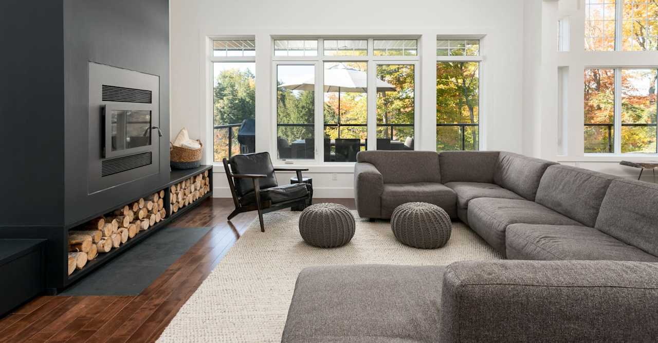 Living room with a gray sectional, black fireplace, stacked firewood, and tall windows overlooking autumn trees.
