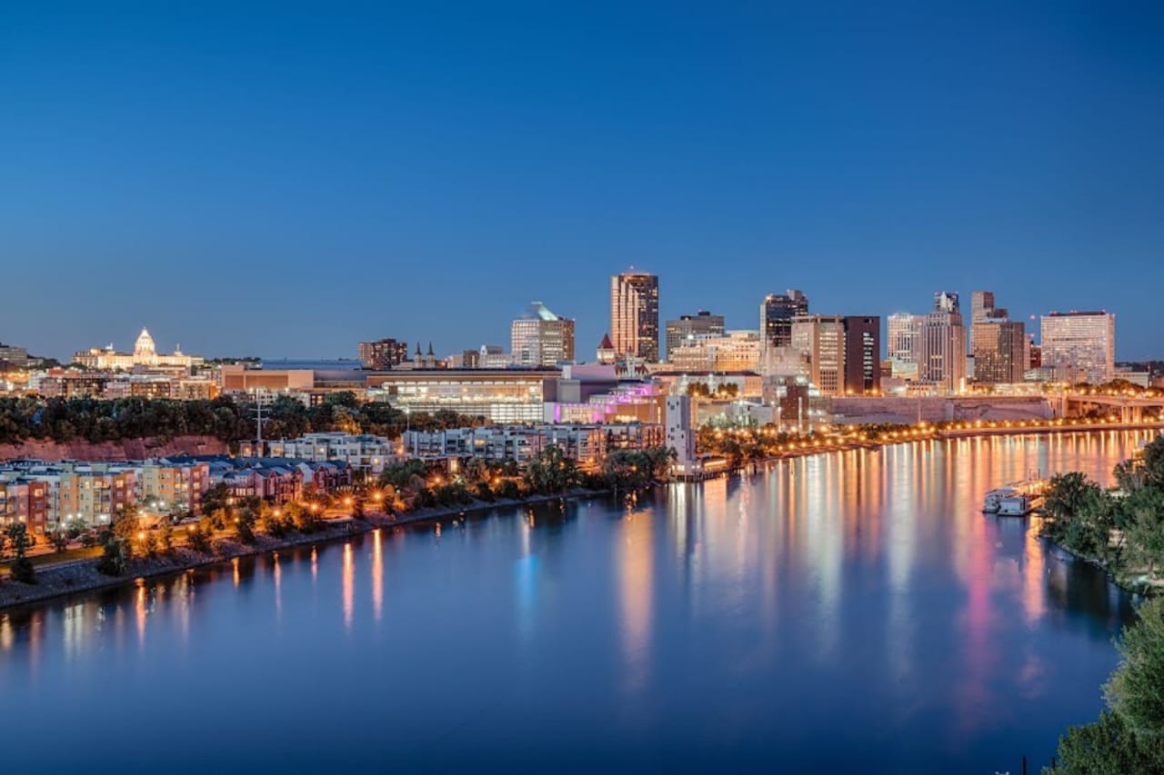 A cloudless blue city skyline with tall and modern buildings, bright lights and a wide and serene river in the foreground.