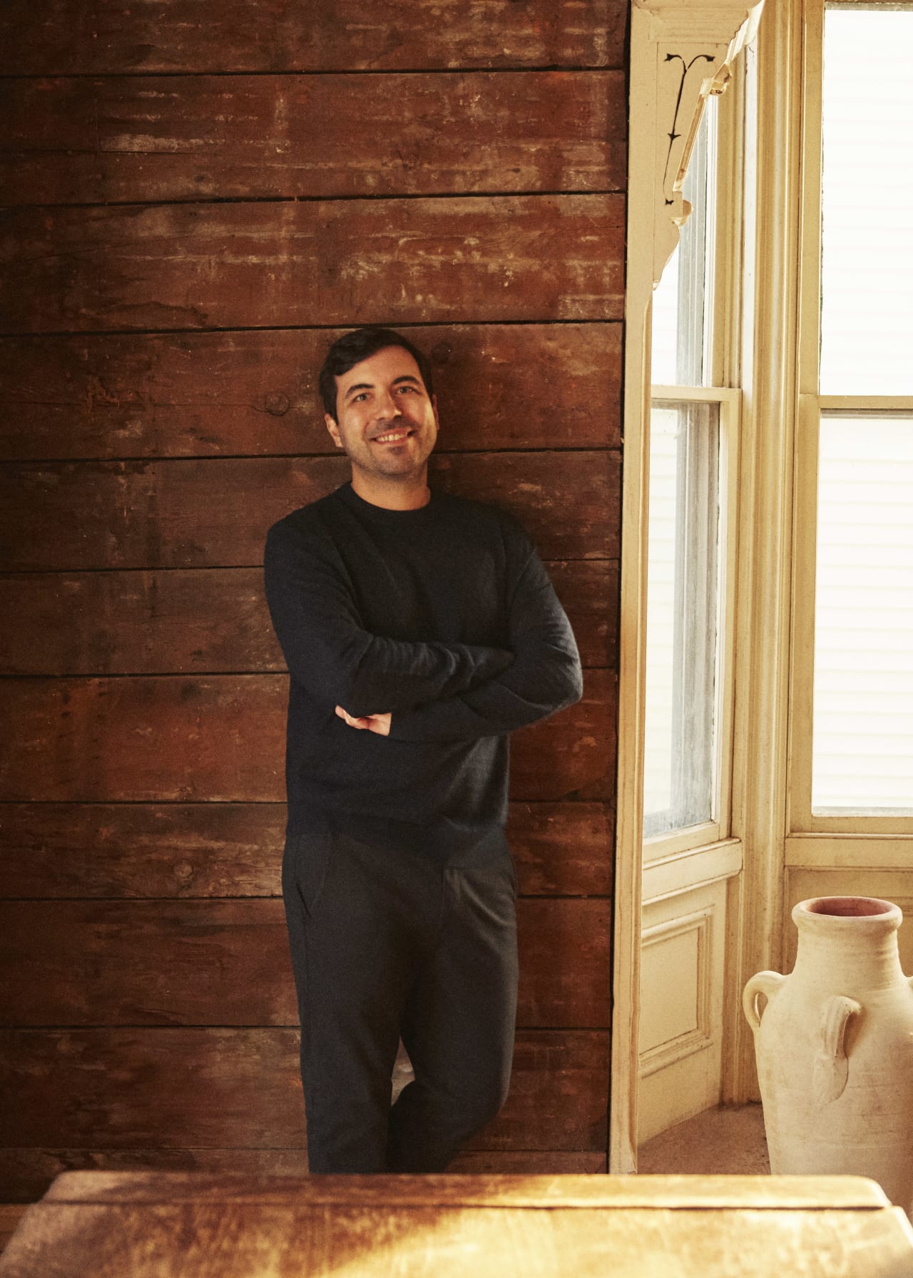 A man with a warm smile, wearing a casual dark blue long-sleeve shirt and black pants, stands leaning against a rustic wooden wall near an open doorway. He has crossed arms and appears relaxed and approachable. The natural light from the adjacent room illuminates one side of his face, highlighting a friendly and confident expression.