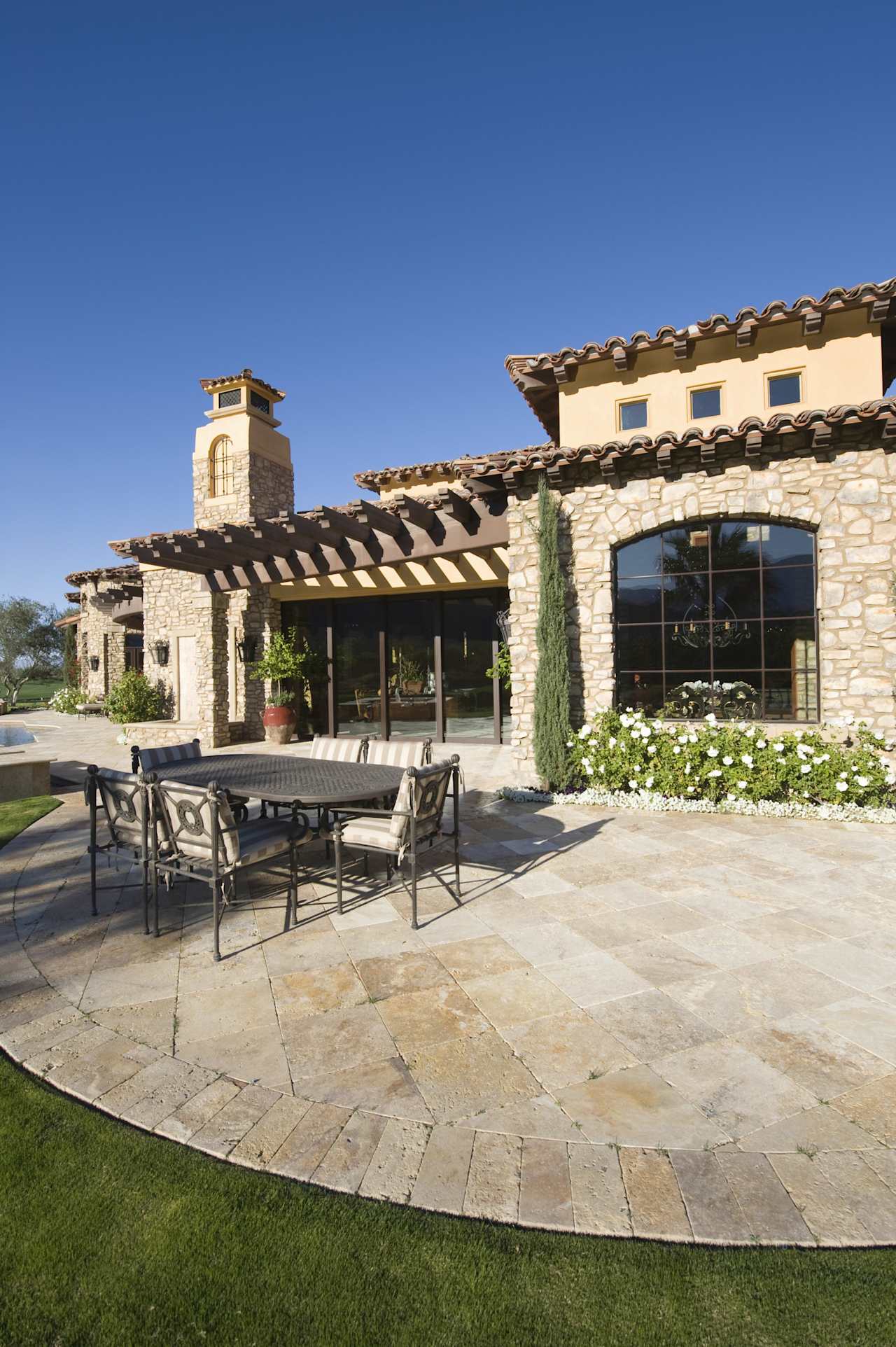 A brown house with a brick patio, a wrought iron table, four chairs in front, and scattered plants on the side.