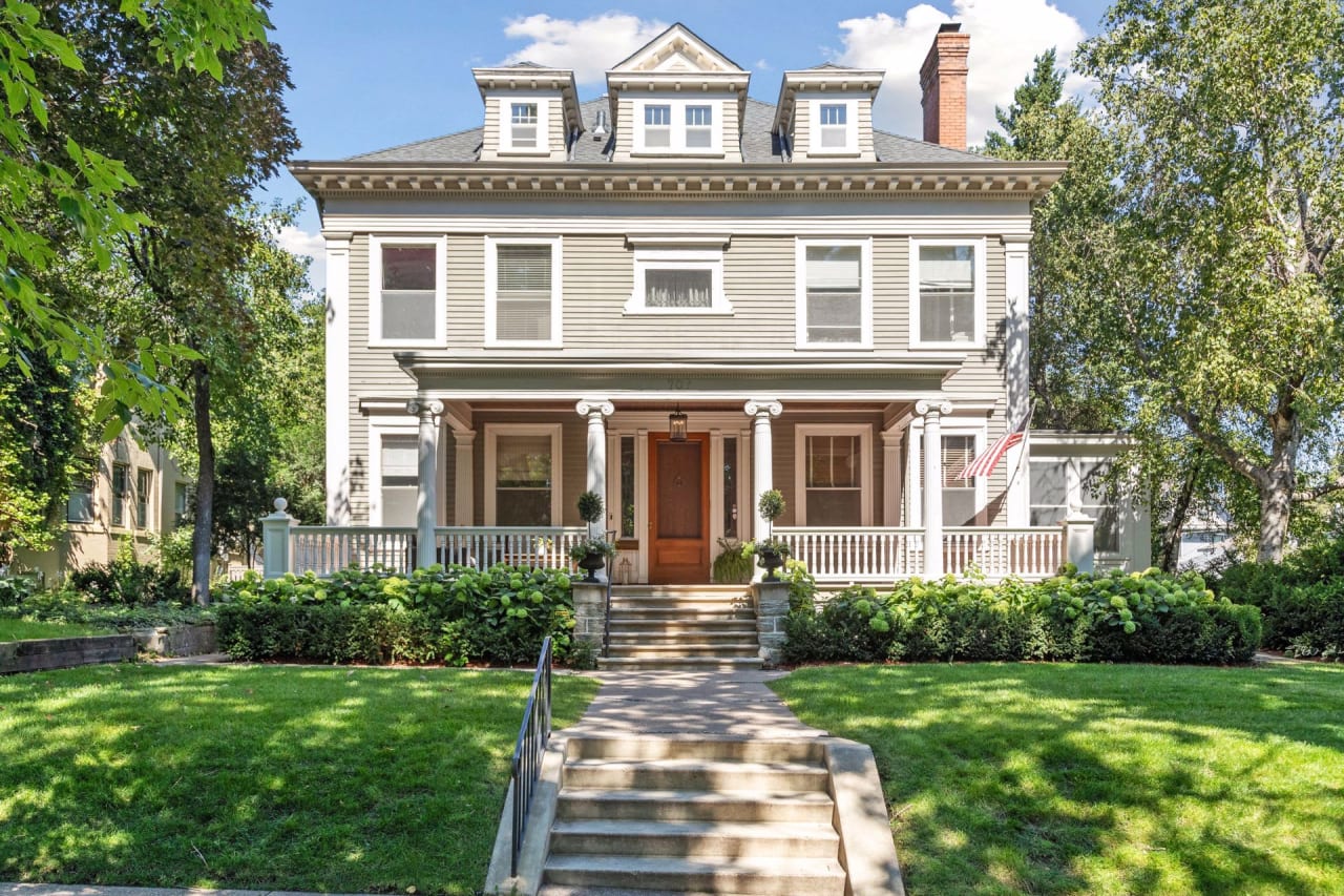 A large gray house with white trim and stairs leading to the front door. Has a wraparound porch with columns and a balcony.