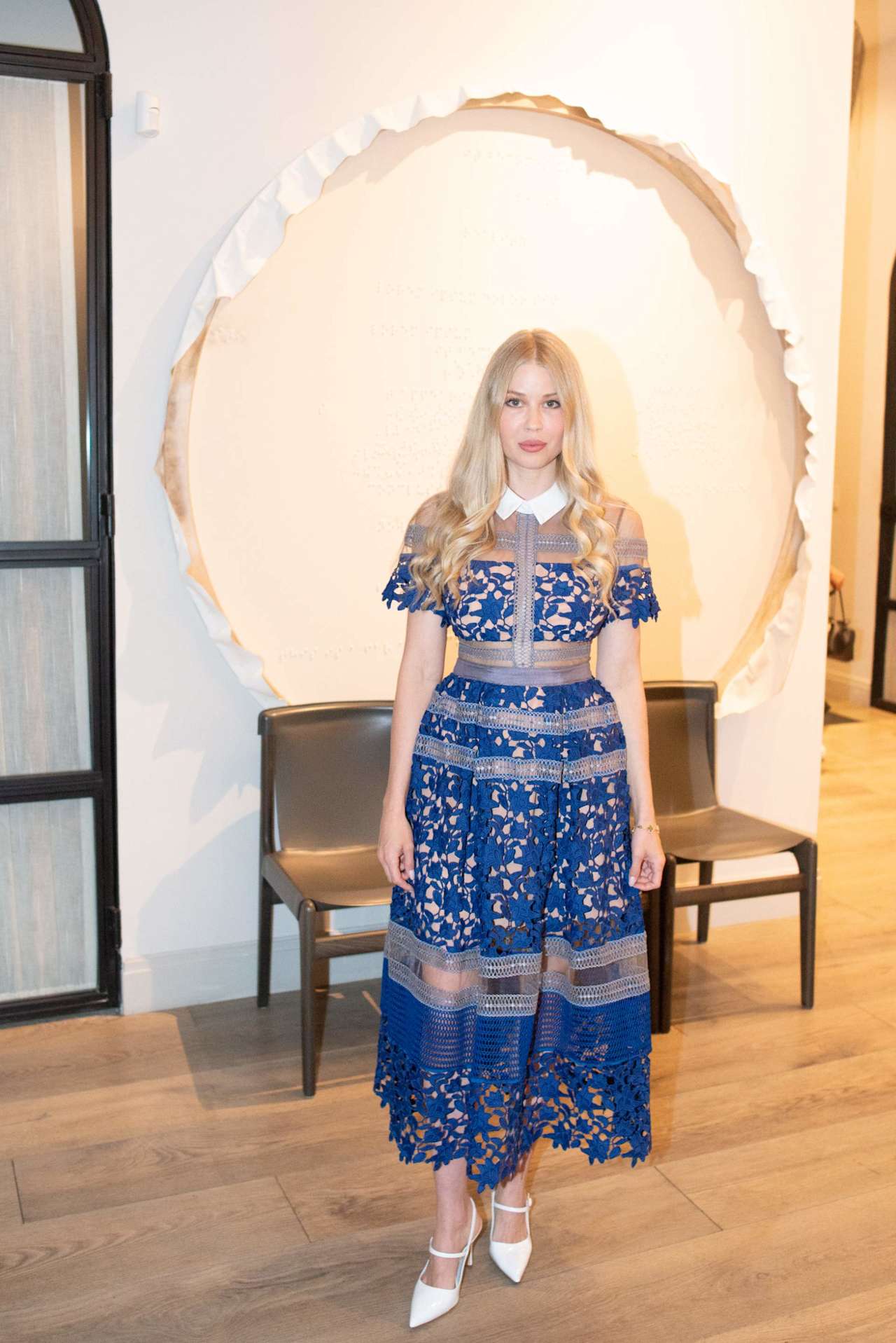 Woman in a blue lace dress posing in front of a large circular wall feature.