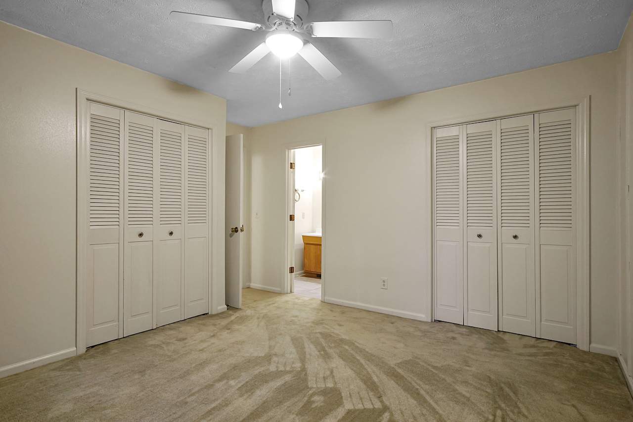 An interior of a bedroom with cream carpeted flooring, cream walls, a ceiling fan, and multiple closet doors.