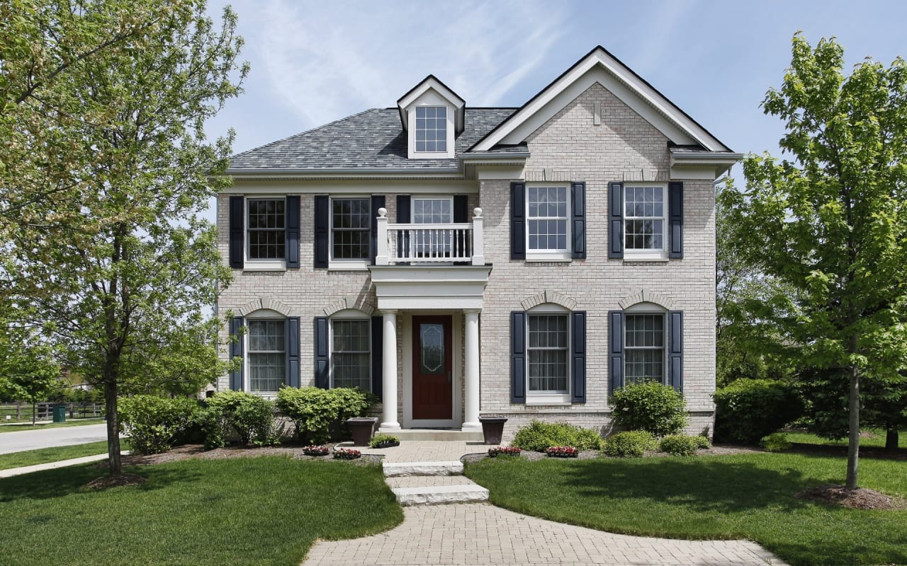 A two-story white brick house with black shutters, a garage, a walkway to the front door, and surrounded by trees and bushes.