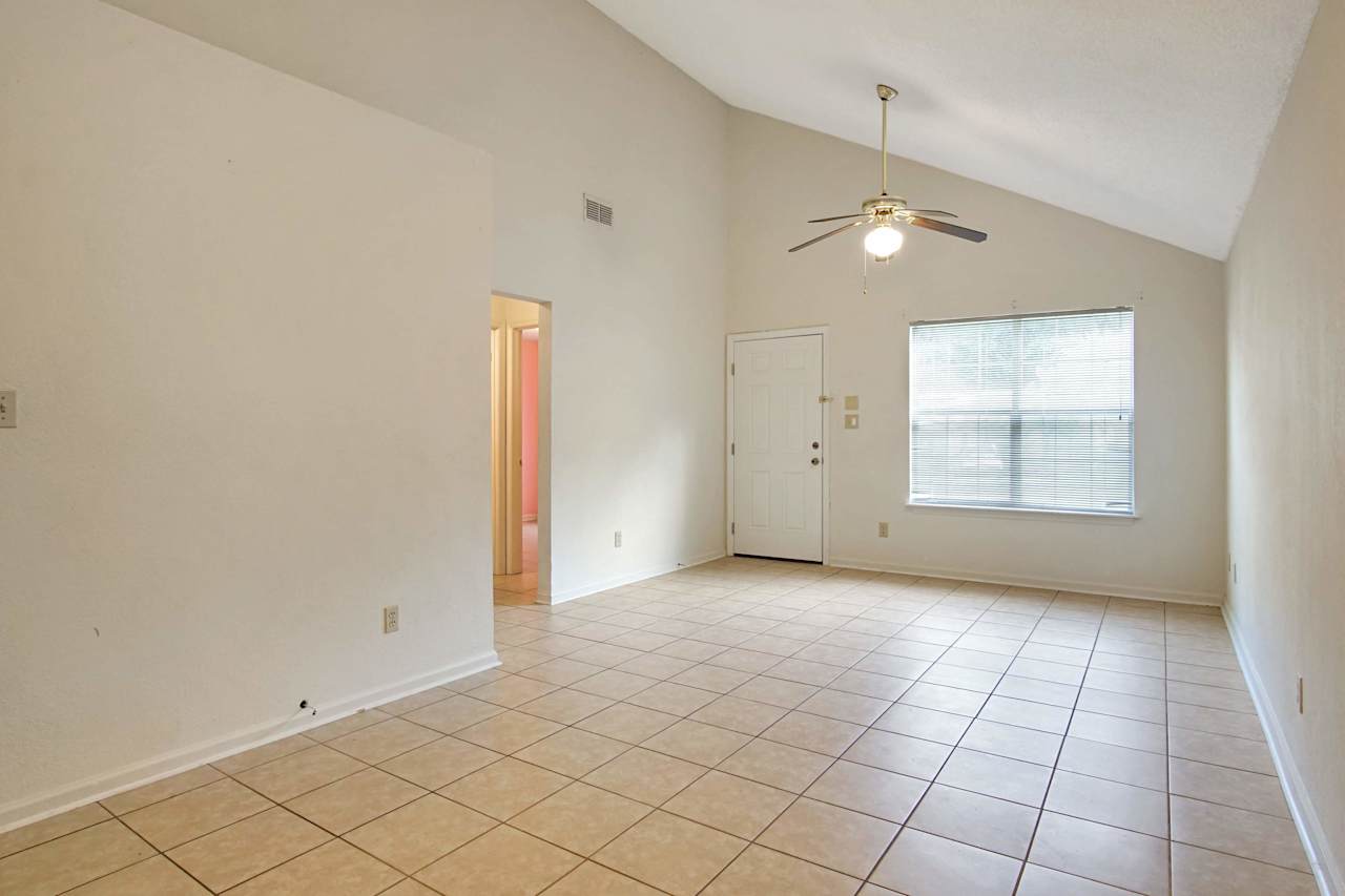 Photo of Maine living area featuring lightly colored walls, bolted ceilings, lightly colored tile, a large window with white pane and white blinds, the white front door entrance, and the hallway to the second and third bedrooms and guest bathroom  at 2709 Oak Park Court, Tallahassee, Florida 32308