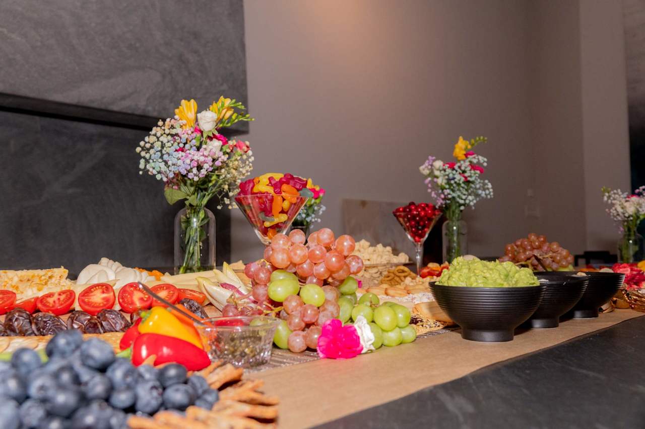 Indoor buffet with grapes, cheese, crackers, vegetables, sweets, and small flower arrangements.