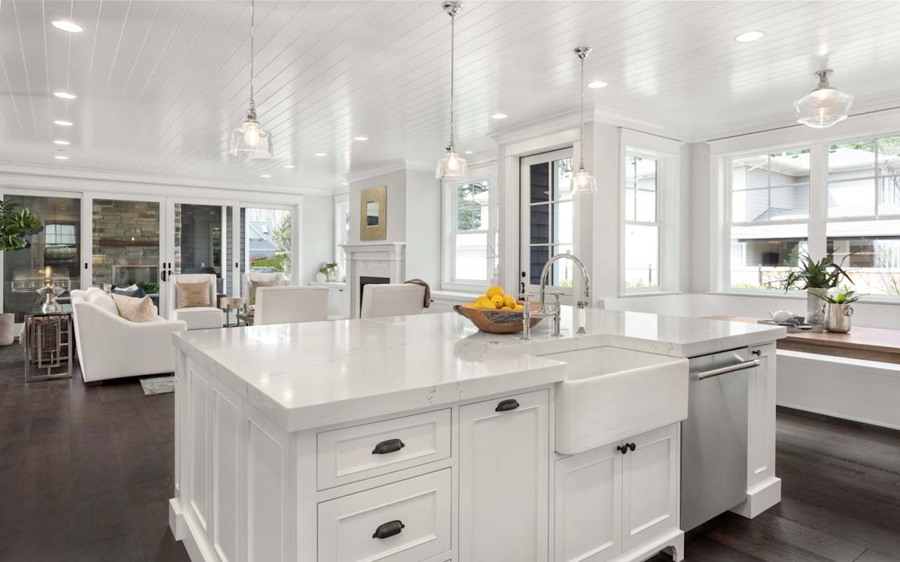 Bright open-plan kitchen with a large white island, farmhouse sink, and windows overlooking the outdoors.