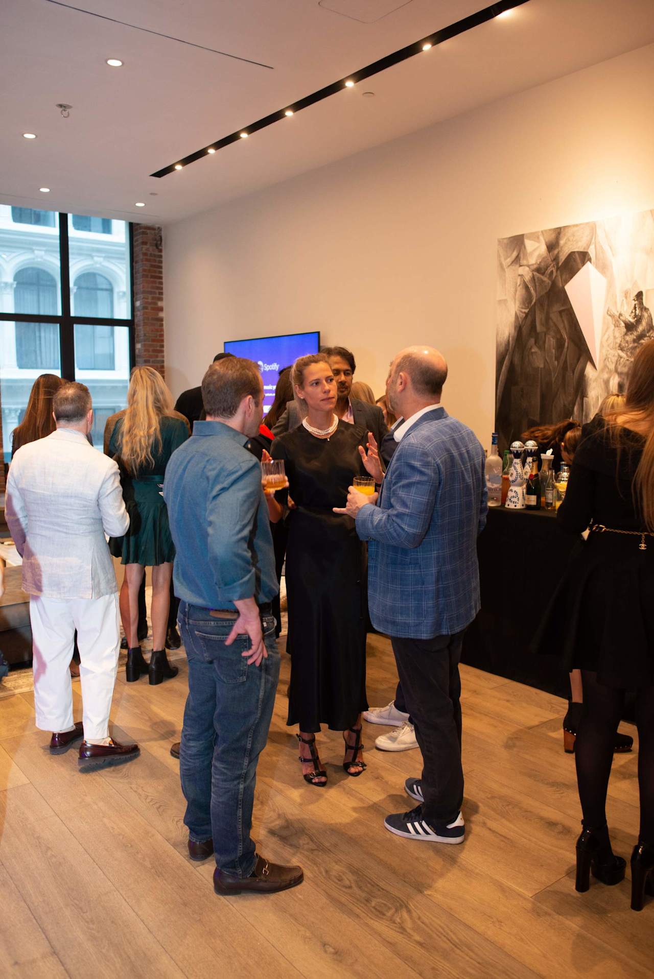 Adults mingle at a company event beside a refreshments table and large windows.
