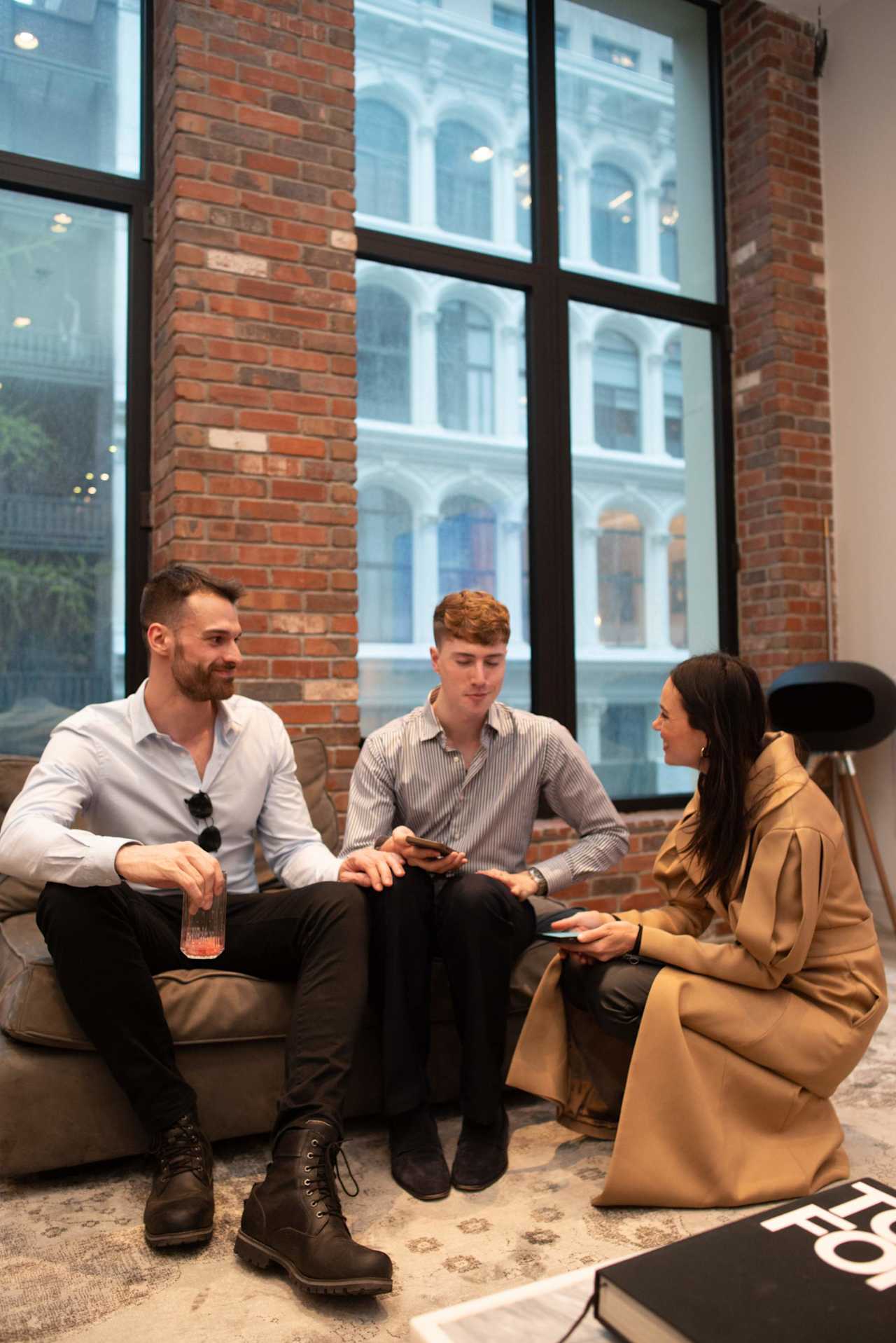 Three people converse on a sofa in a brick-walled office lounge with tall windows.