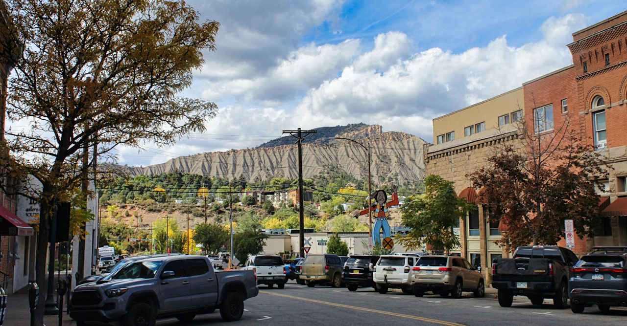 Downtown street with brick storefronts and parked vehicles beneath a dramatic striated mountain ridge.