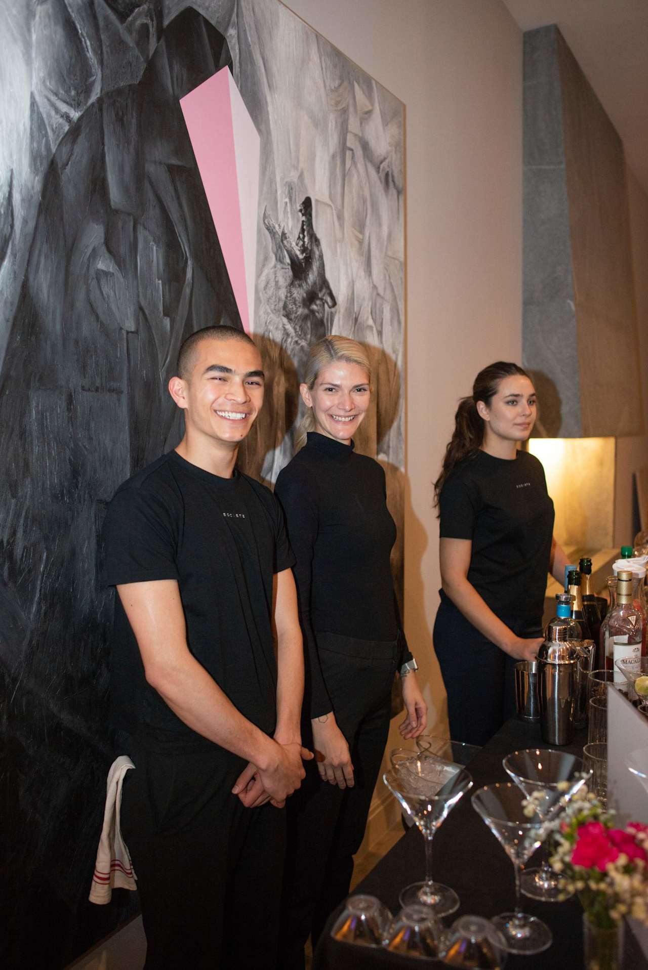Three people in black stand beside a bar with bottles and glassware.