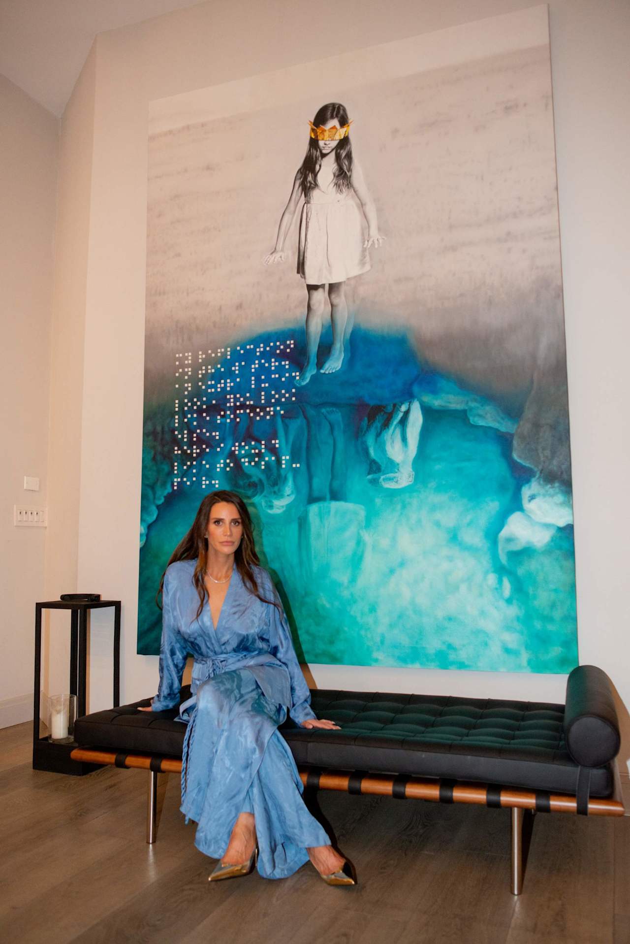 Woman in a blue dress seated on a black sofa before a large turquoise artwork.