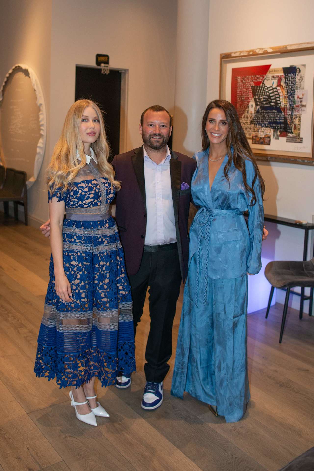 Three adults pose together indoors, with the women wearing blue dresses.