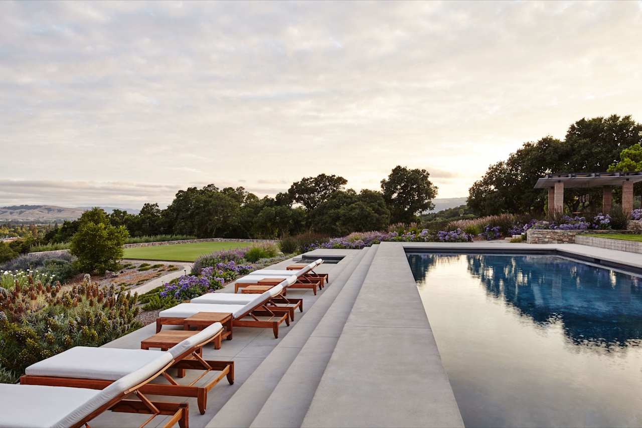 A row of wooden lounge chairs next to a swimming pool. There are trees and plants surrounding the pool area.
