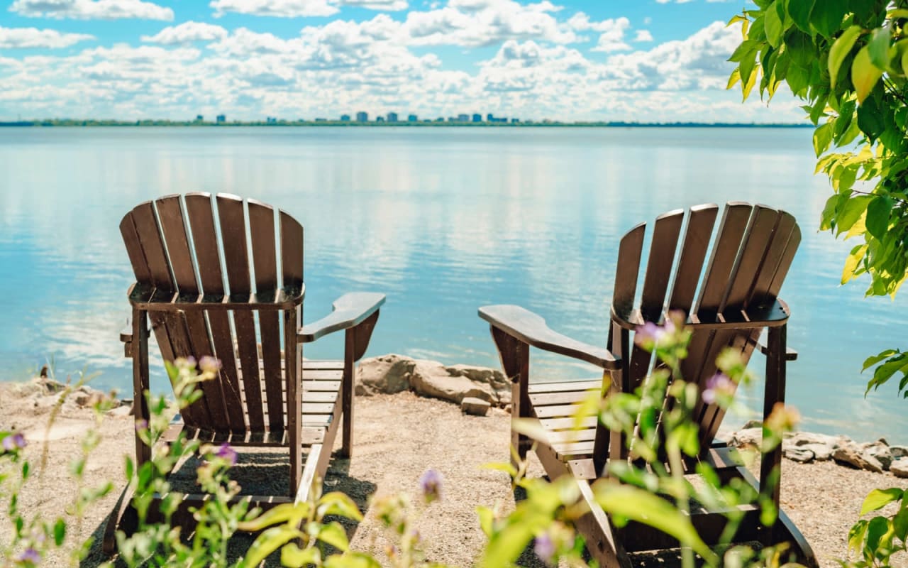 Two lawn chairs sitting on the shore of a lake, with a mountain range in the background.