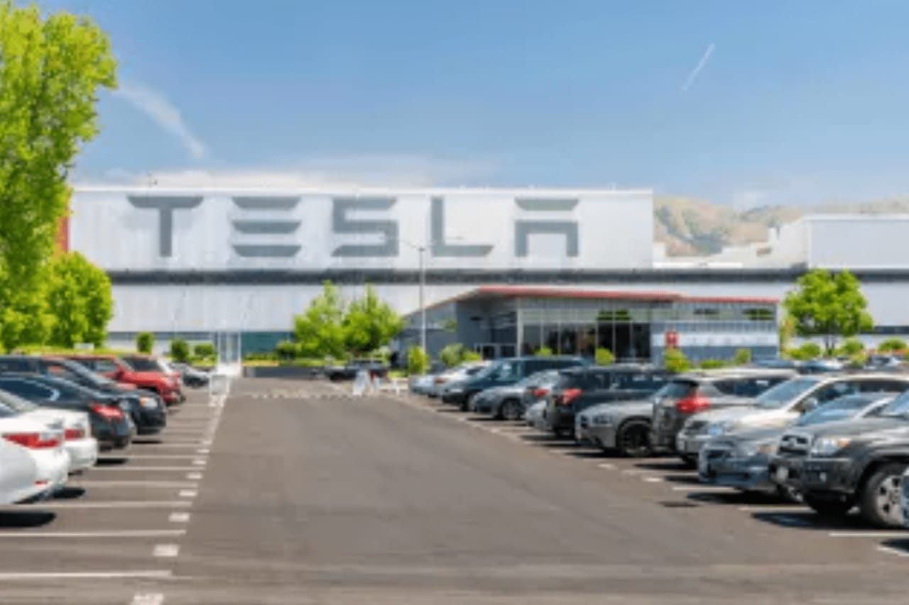 The image shows a parking lot in front of a Tesla factory. There are many cars parked in the lot.