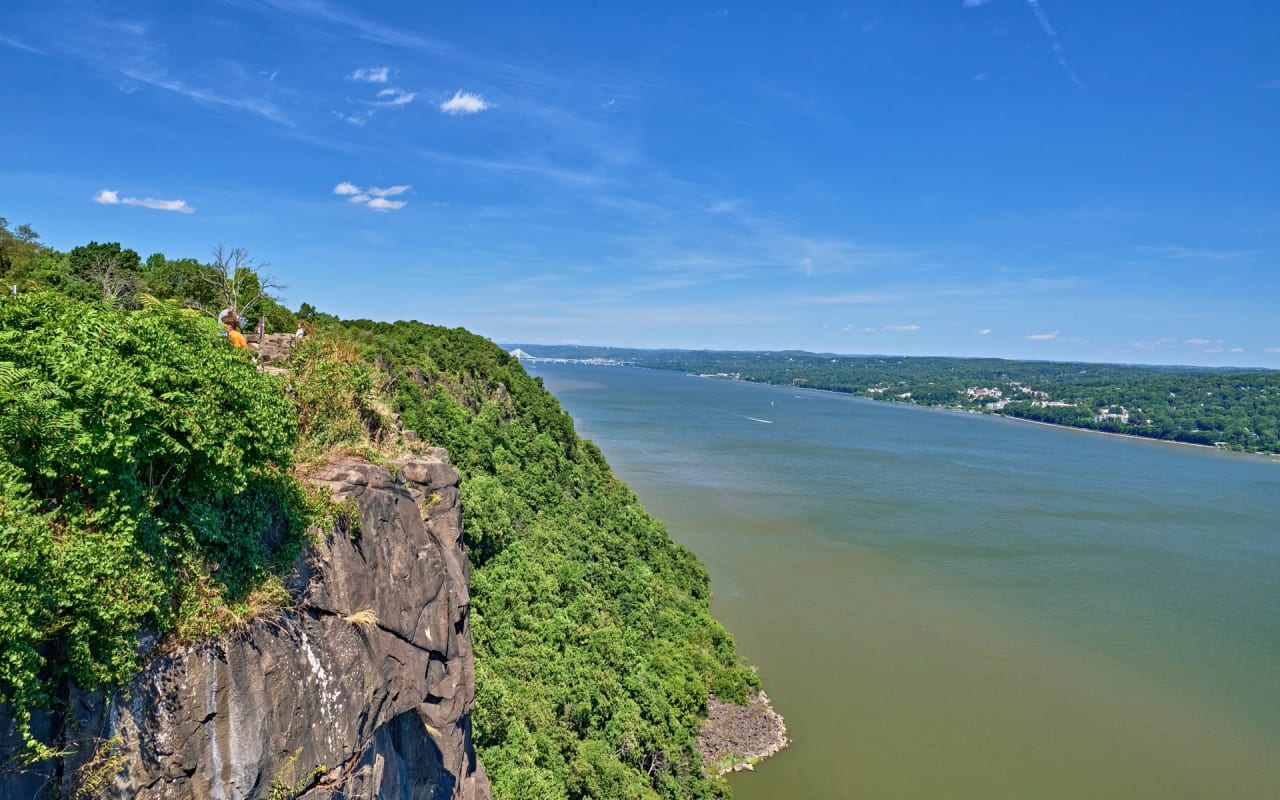 A rocky cliff covered in trees and vegetation, overlooking a view of winding river and distant hills in Palisades Interstate Park, New Jersey.