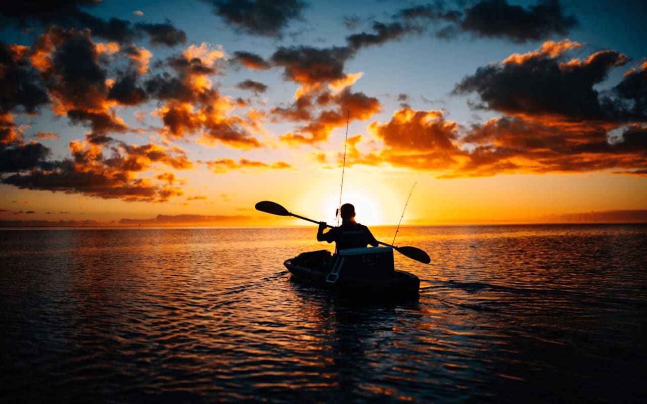 Silhouetted kayaker casts a line as the sun dips below the horizon, painting the sky in fiery hues.