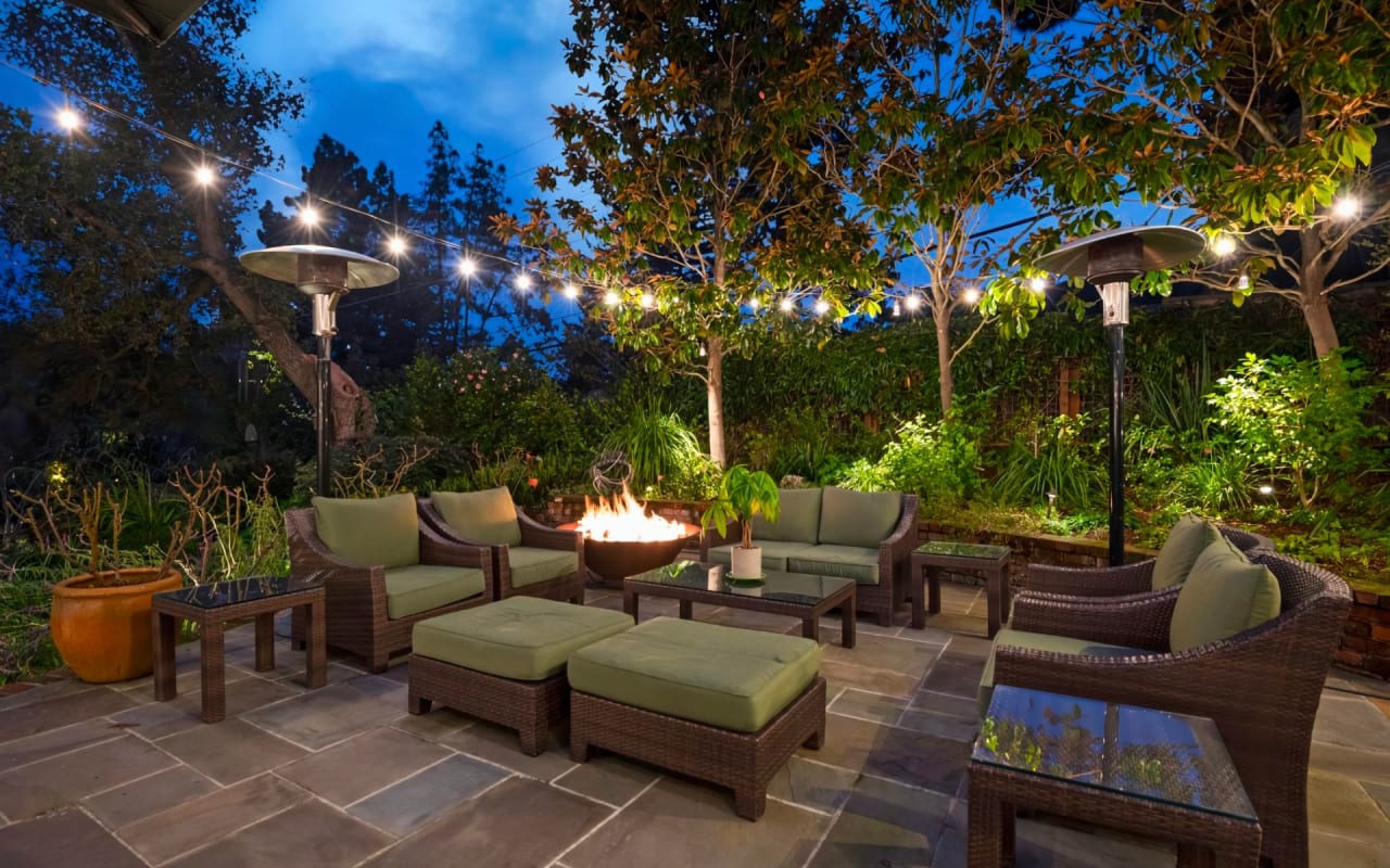 A patio with wicker furniture, a round fire pit centerpiece, and several trees and other plants around the perimeter.