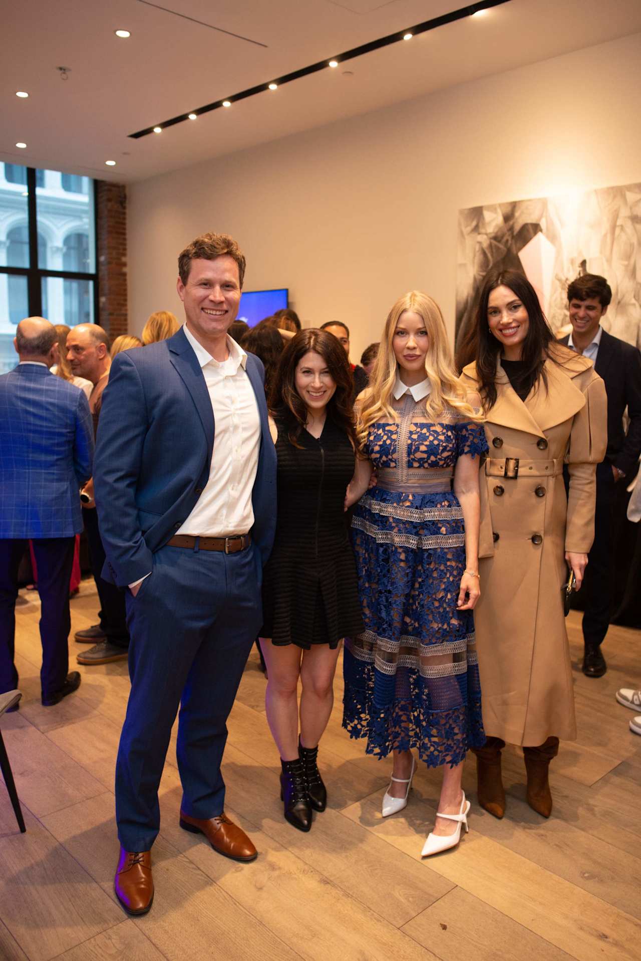 Four event attendees pose together in a crowded venue, dressed in a blue suit, blue lace dress, and tan coat.