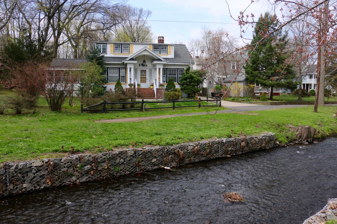 A large white house with a black roof, chimney, and basement, next to a stream and surrounded by greenery.