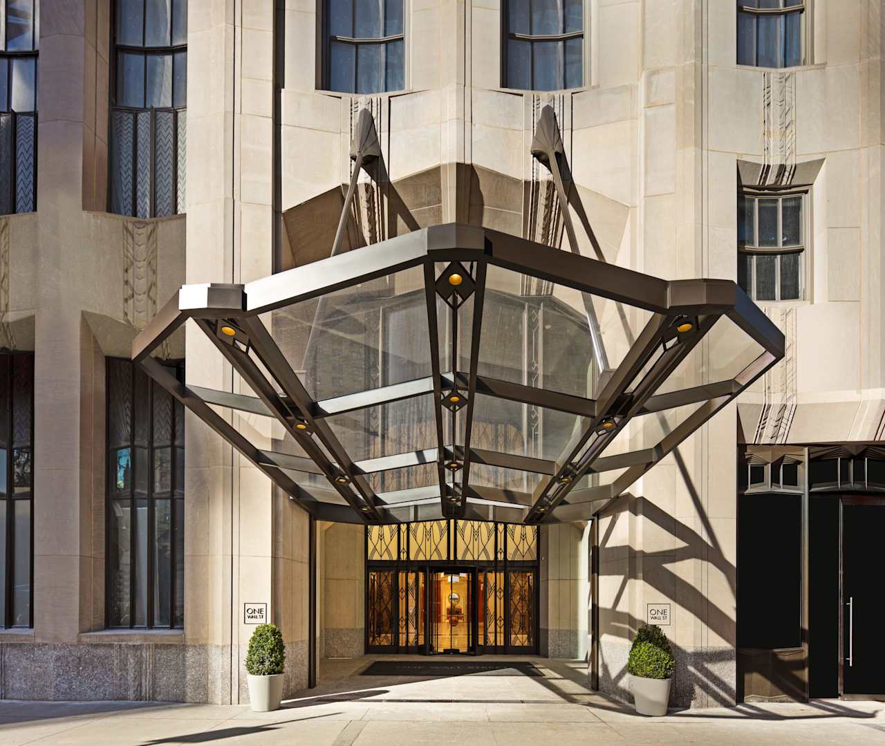 A modern hotel entrance featuring a glass canopy, elegant lighting, and decorative planters. The facade showcases architectural details.