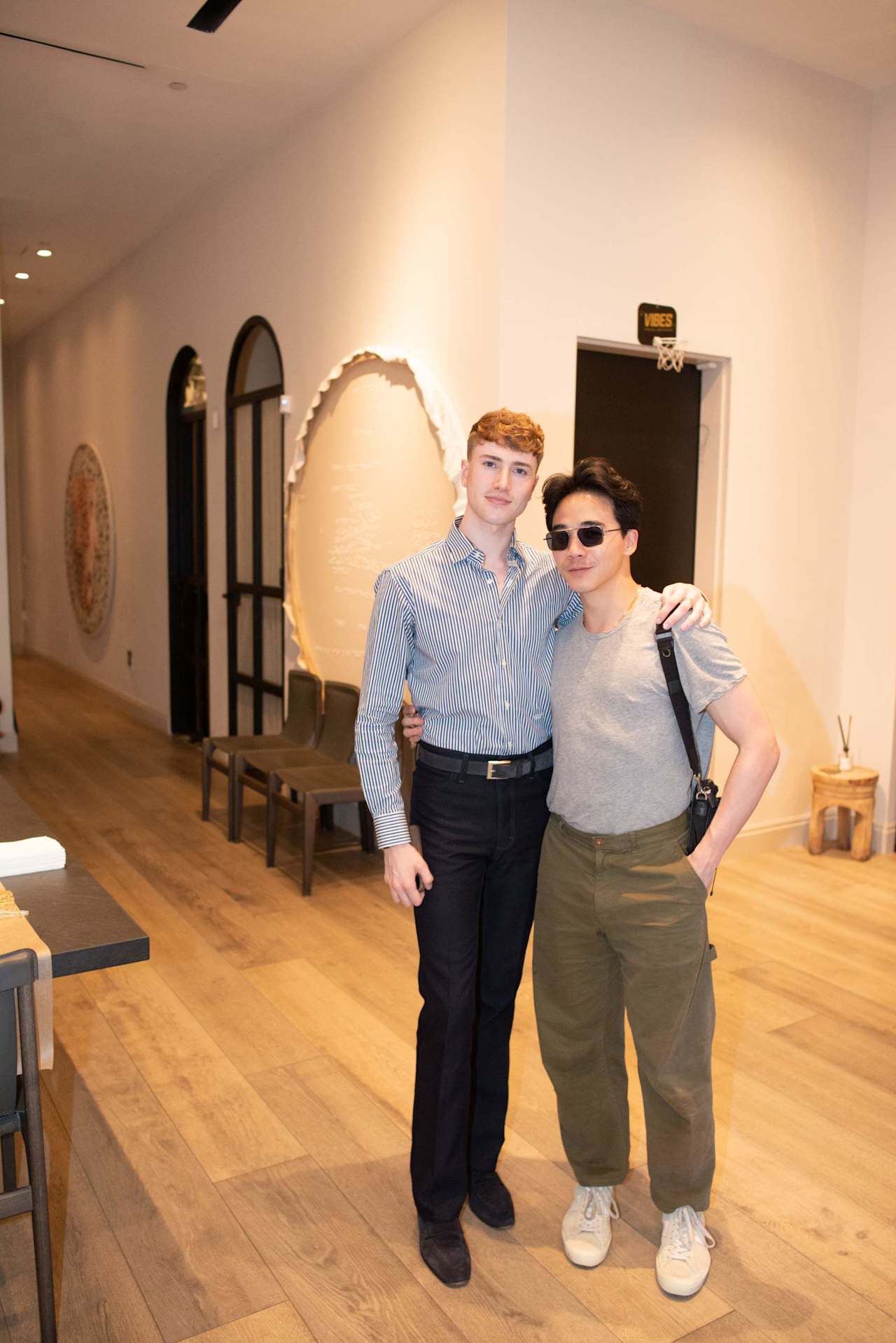 Two men pose together in a real estate office, one wearing a striped shirt and the other sunglasses.
