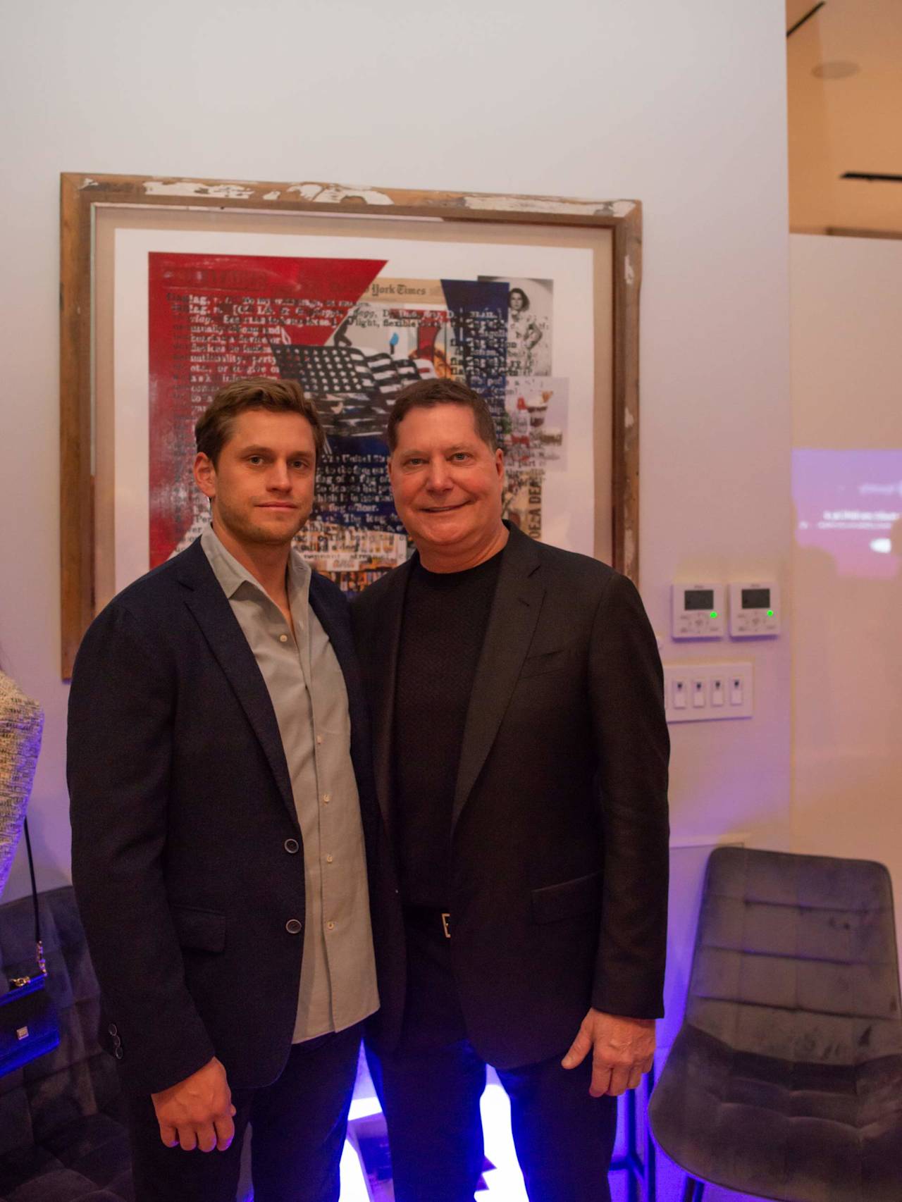 Two men pose in dark jackets before a large framed artwork.