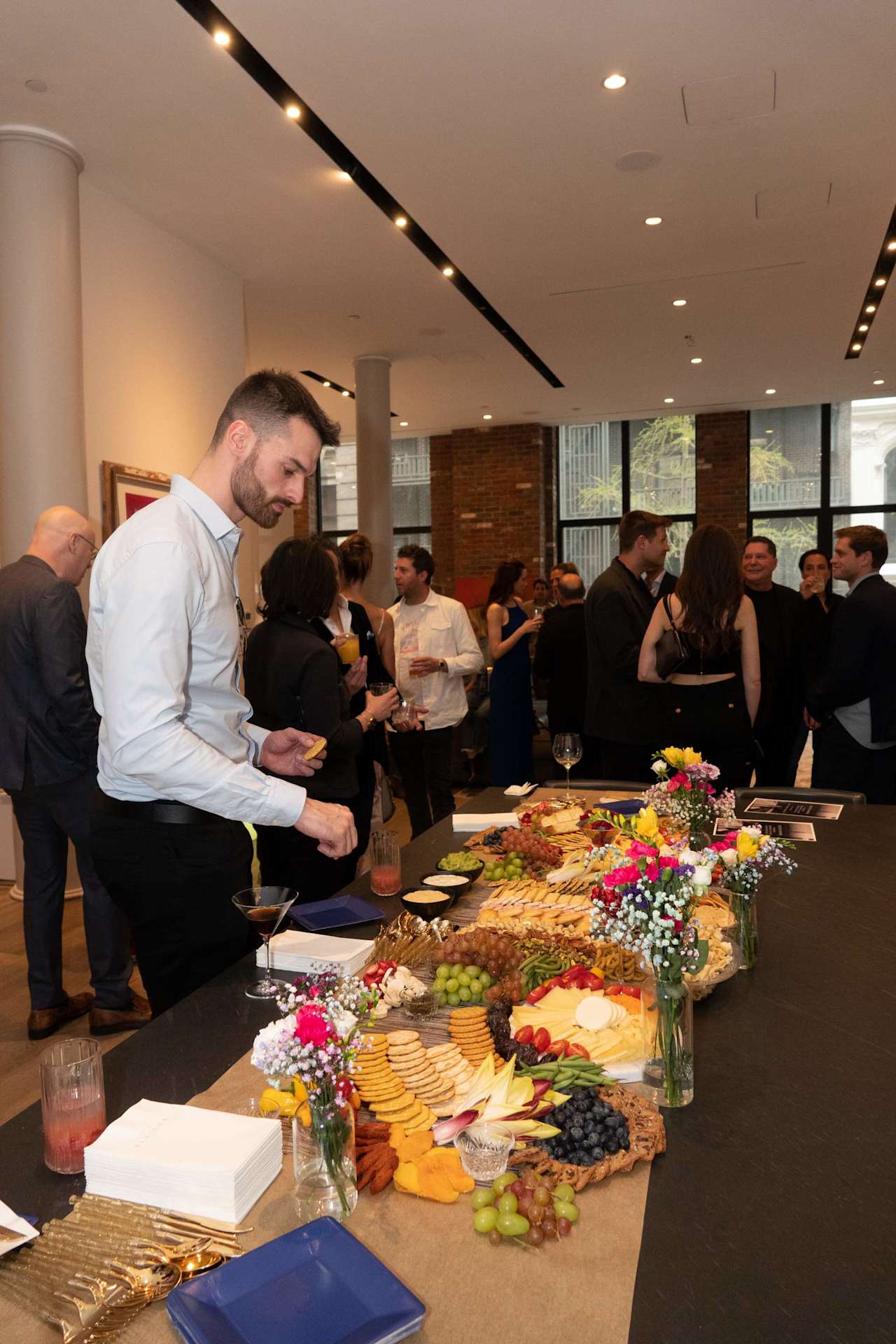 Guests mingle beside a buffet spread with fruit, appetizers, and floral arrangements.