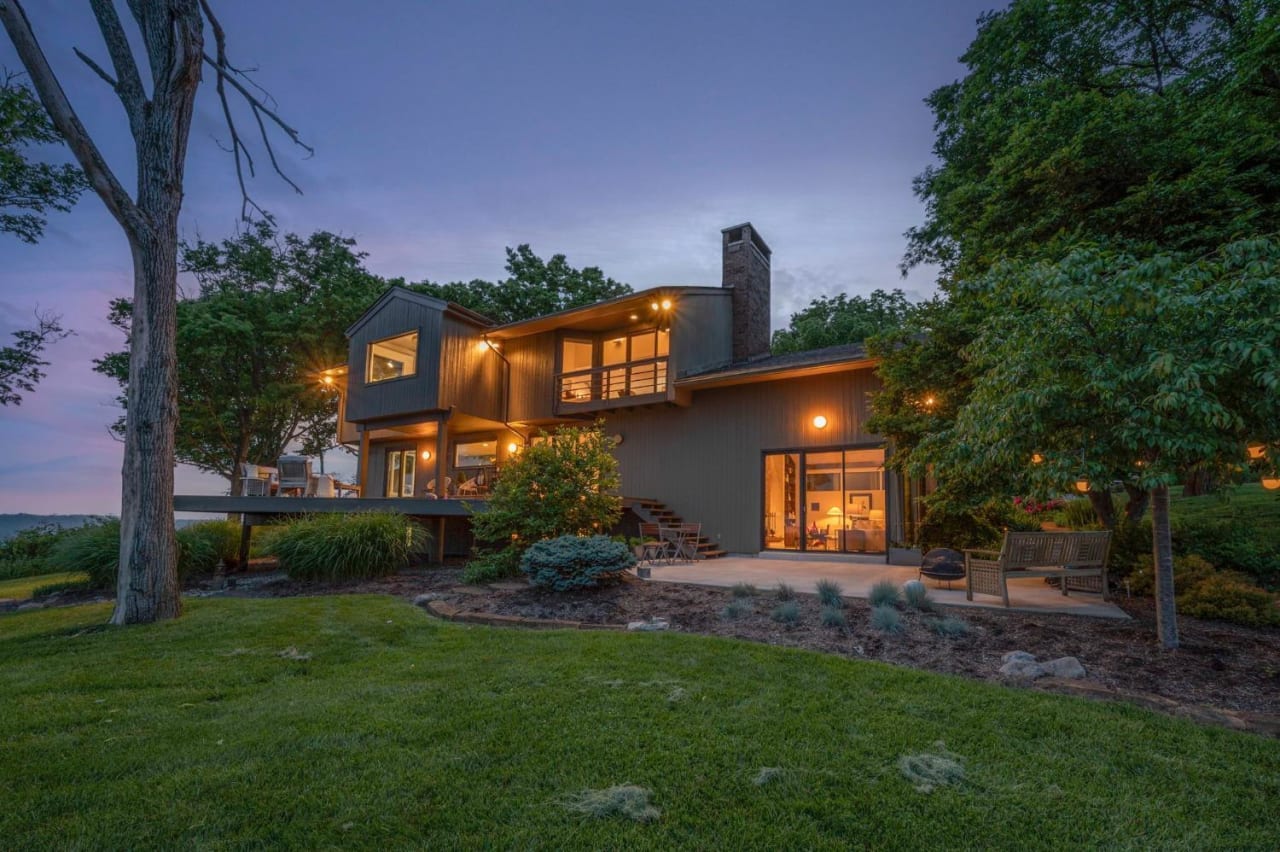 A large house on a hilltop, surrounded by trees. The house has a deck and a fireplace, and there's a large lawn in front