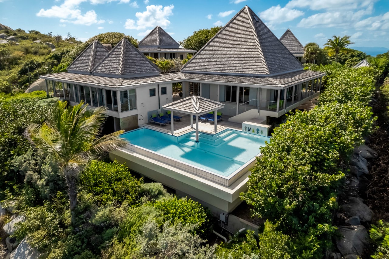 An aerial view of a villa with a large pool, located in a green area with palm trees