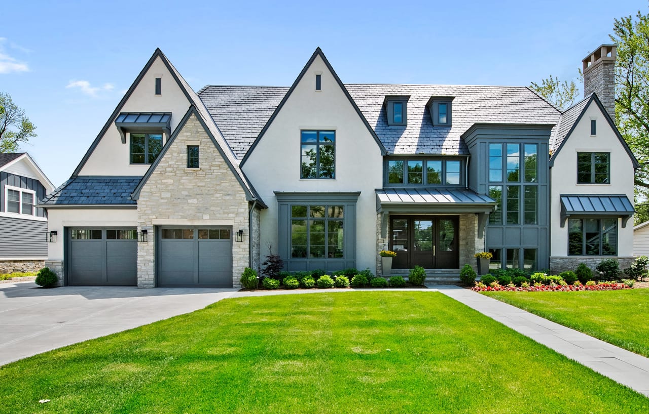 Modern two-story house with a mix of siding materials, large windows, flat roof, small porch, front yard, and two-car garage.