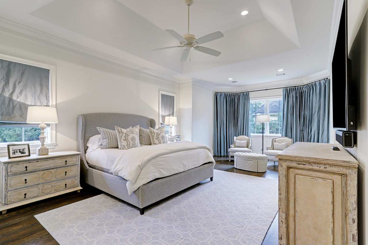 A bedroom with a queen-sized bed, dresser, ceiling fan, a rug, wooden floor, upholstered chairs, and overlooking windows.