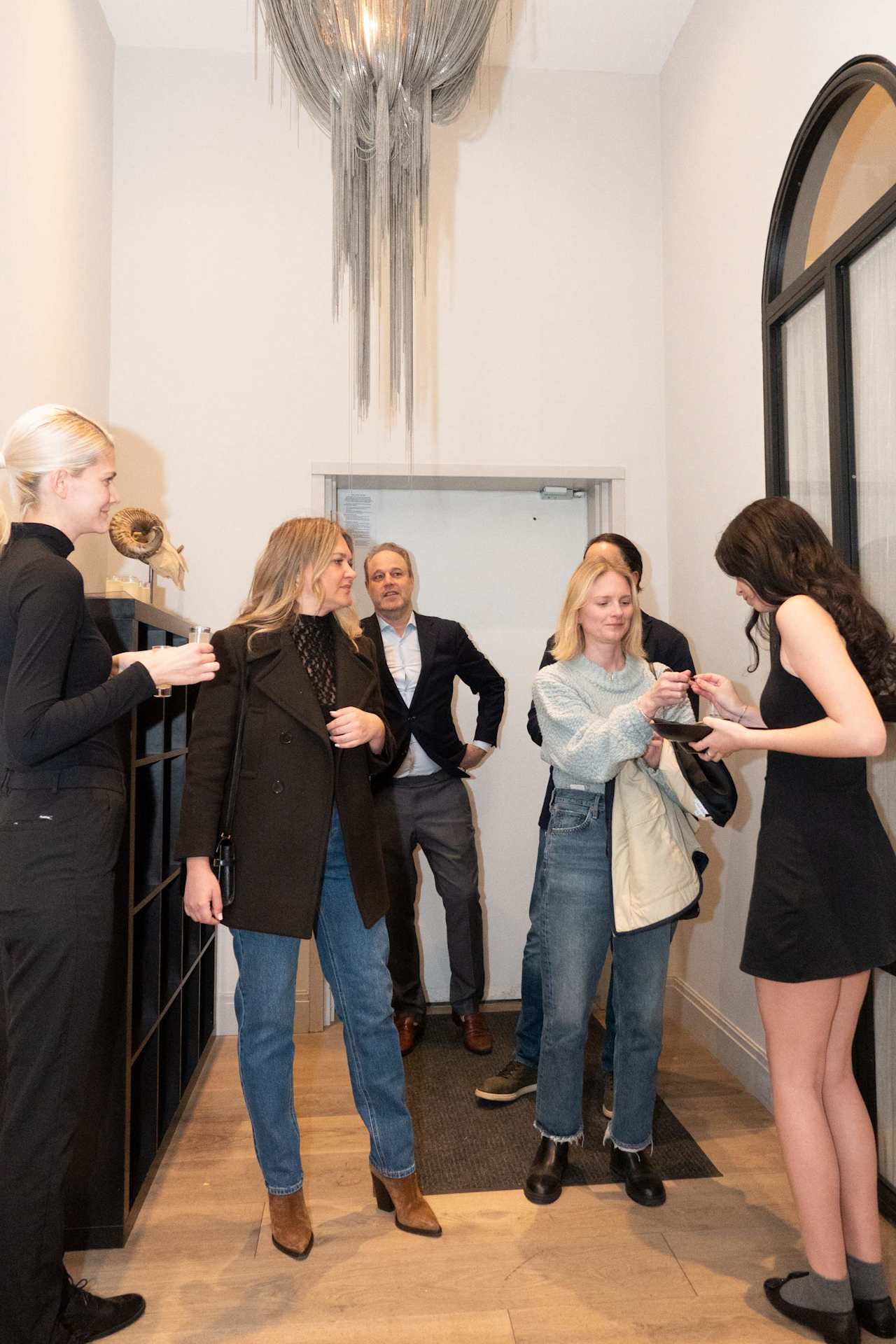 Guests mingle at an indoor gathering beneath a tall chandelier; one serves a small dish.