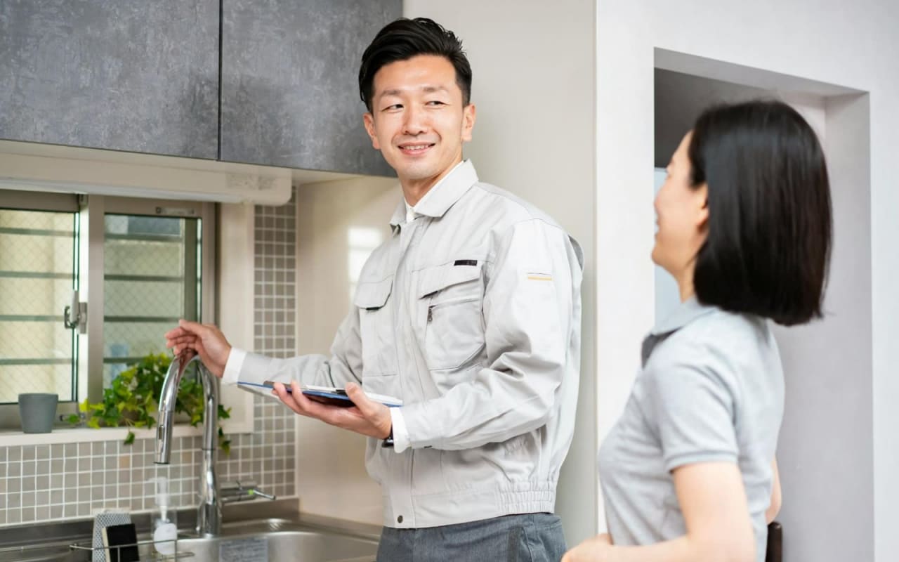 Smiling technician in a gray uniform assisting a homeowner in a kitchen, testing the faucet and holding a tablet.