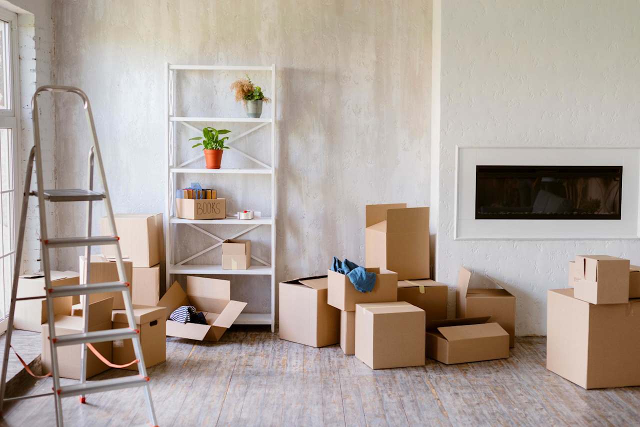 Empty room filled with cardboard boxes, a leaning ladder, and a minimalist bookshelf.