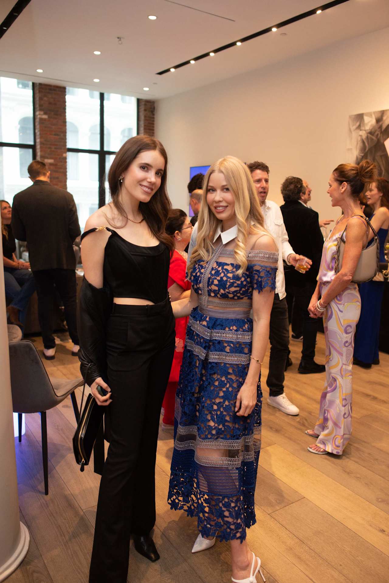 Two women pose at an indoor gathering, one in black and the other in a blue lace dress.
