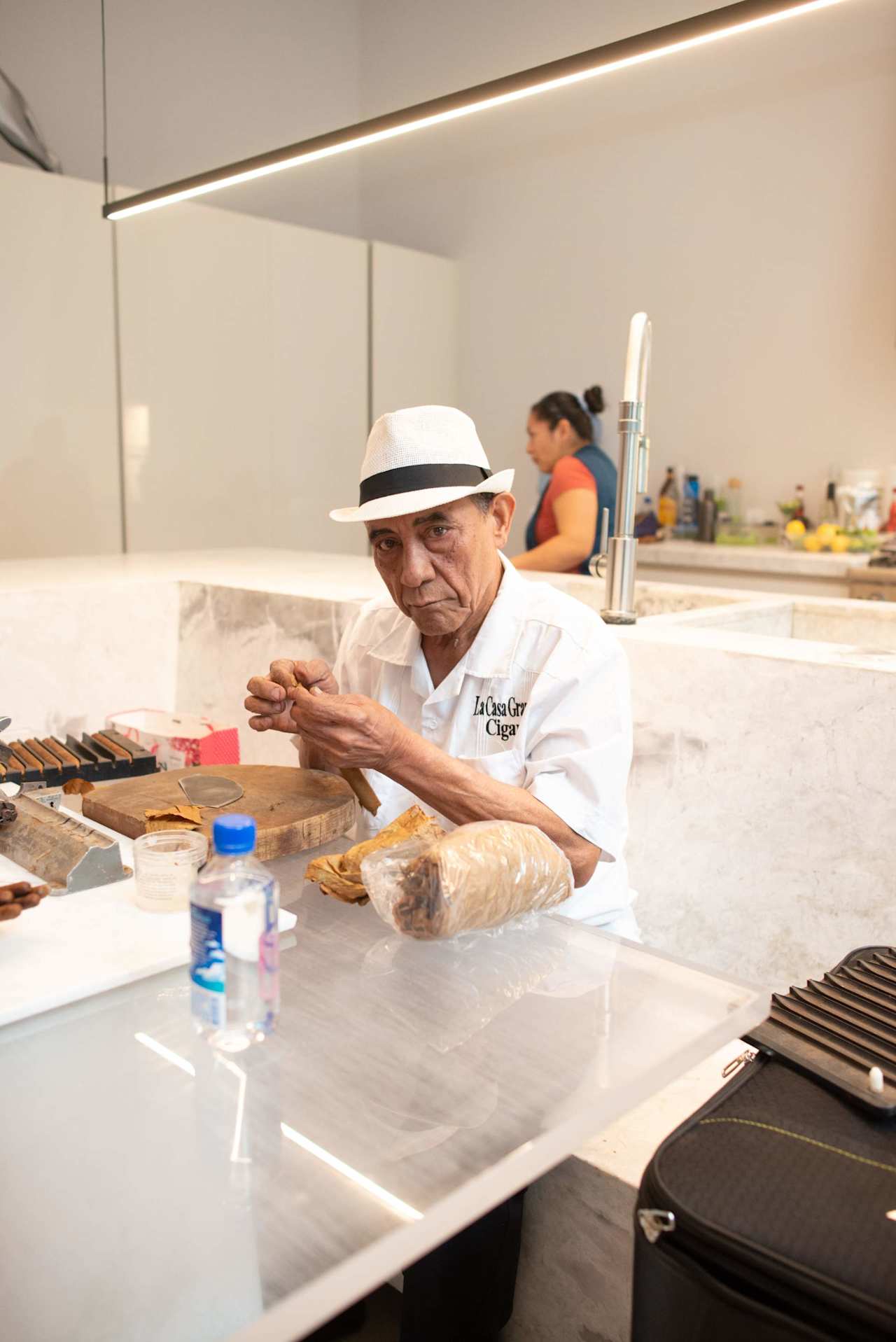 Cigar maker in a white hat works with cigar leaves at a shop counter.
