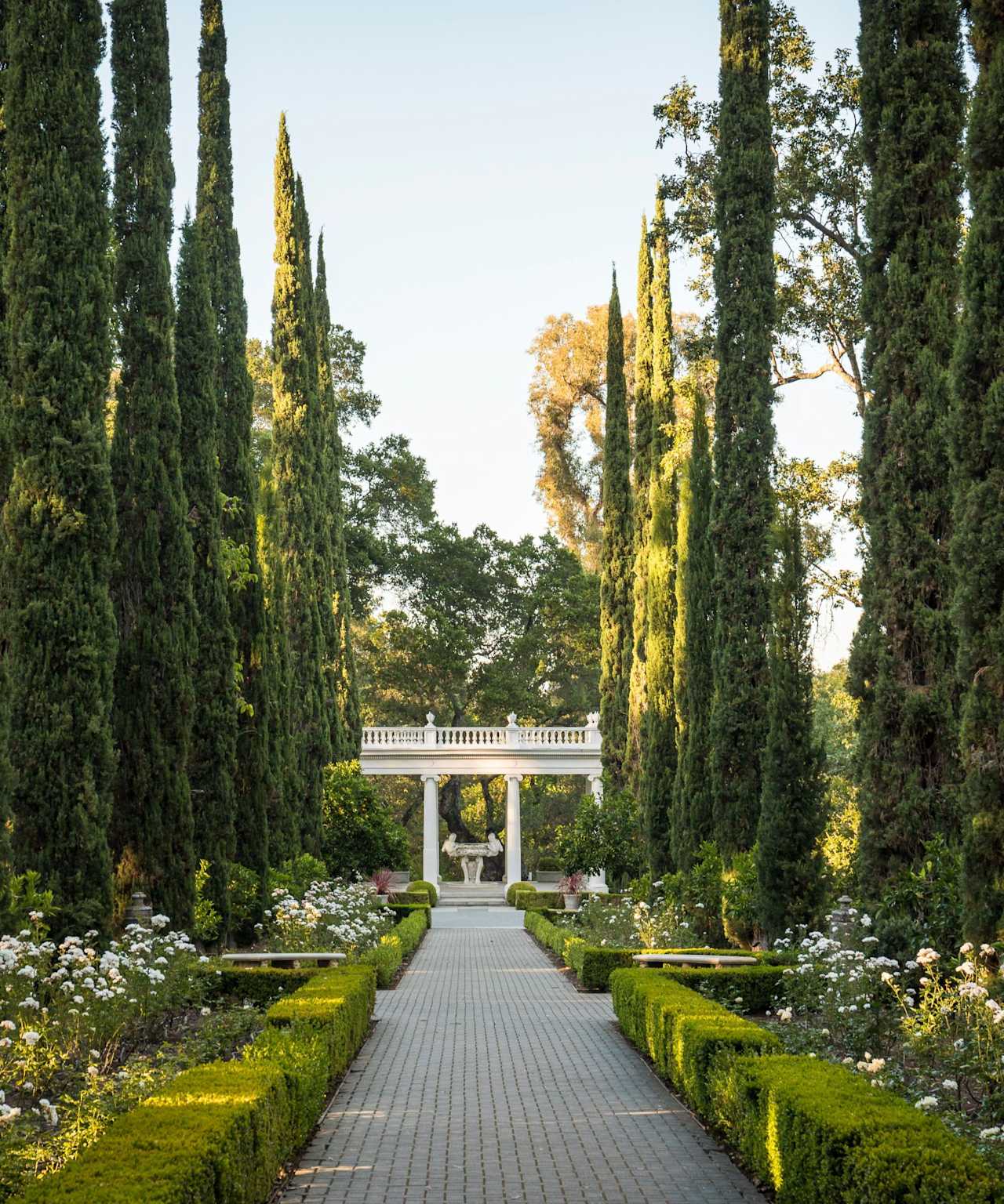 A brick walkway lined with trees and flowers leading to a white gazebo in the background.