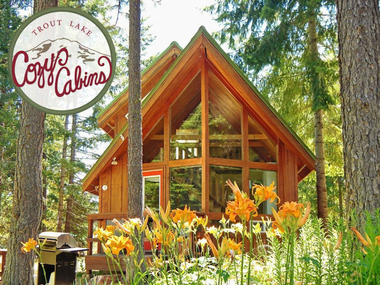 Wooden A-frame cabin among trees and orange lilies, with a Trout Lake Cozy Cabins sign.