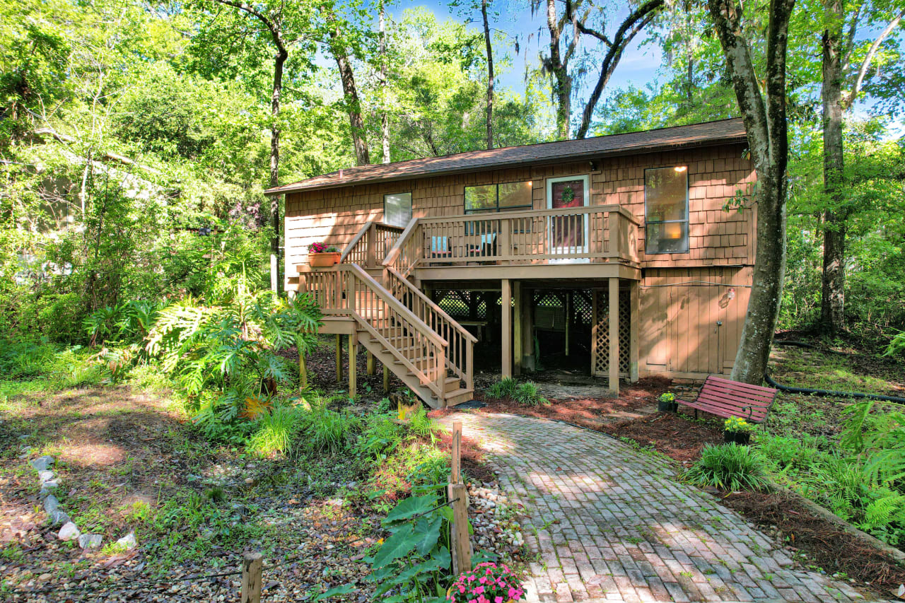 The exterior of a house with a combination of brick and siding. The house is elevated with steps leading up to the entrance and a small deck area. The yard is lush with greenery and trees, creating a serene environment.