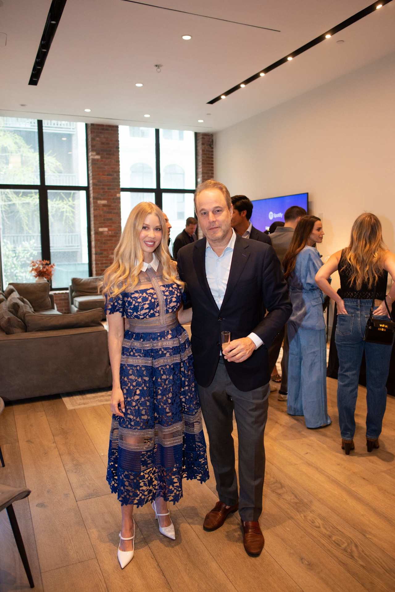 A couple pose at an indoor gathering, the woman in a blue lace dress and the man holding a drink.