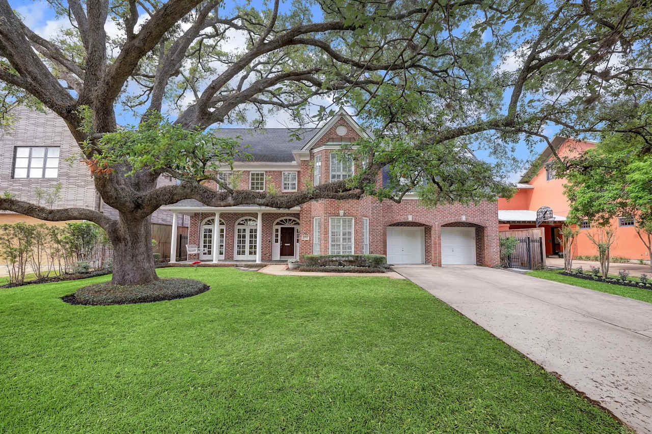 A two-story red brick house with a large front yard, a porch, a paved walkway, and a two-car garage in a quiet neighborhood.