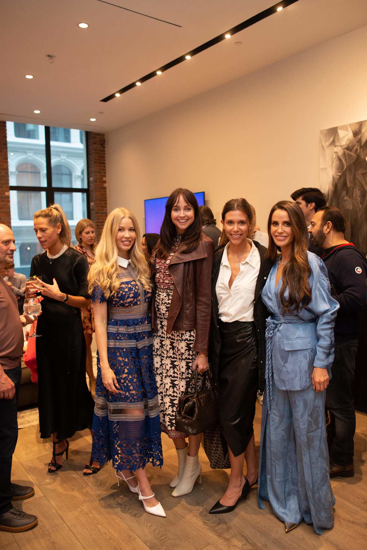 A group of adults pose at an indoor gathering, including women in blue dresses and a light blue suit.