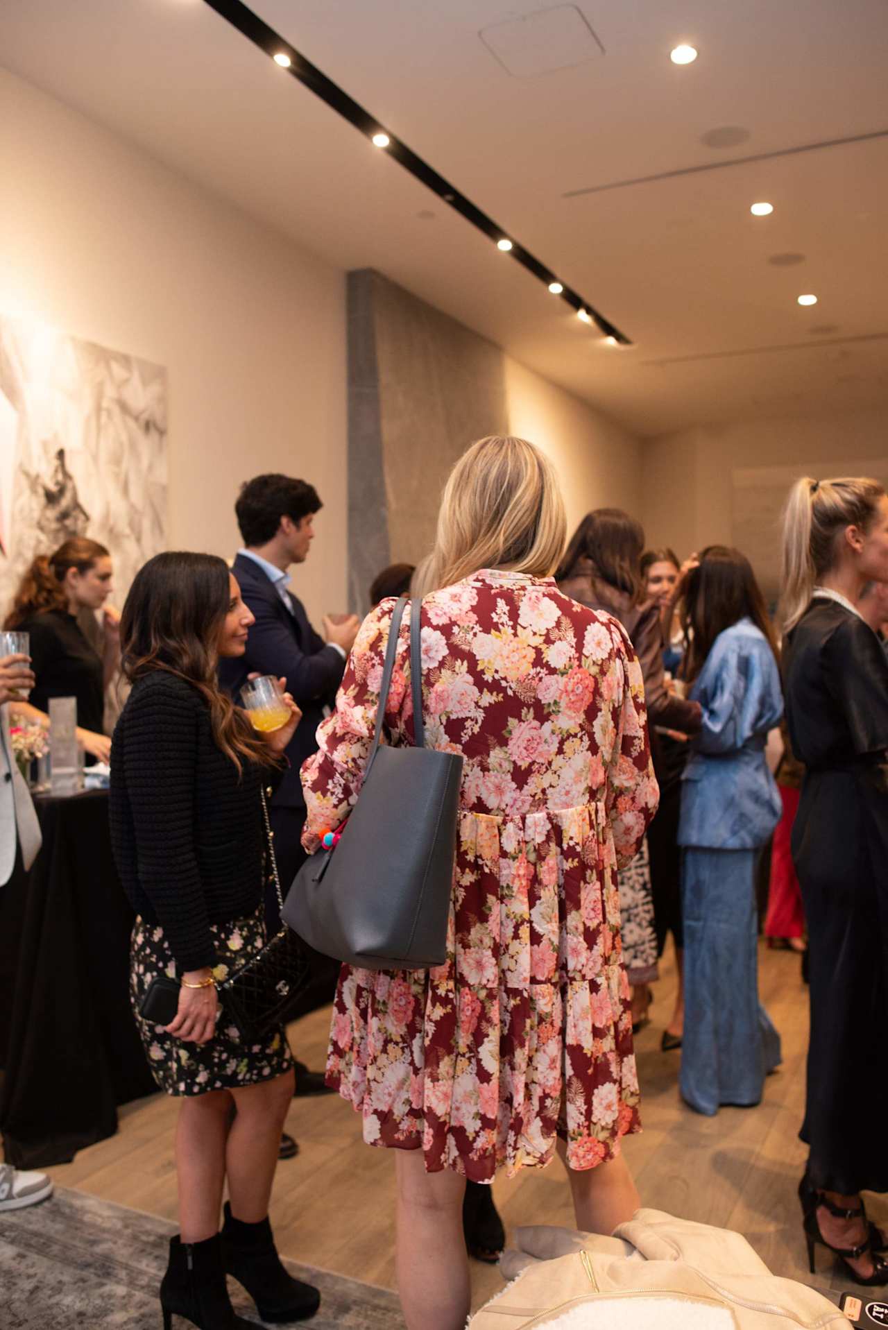 Guests mingle at an indoor event; a woman in a floral dress and gray shoulder bag stands in the foreground.