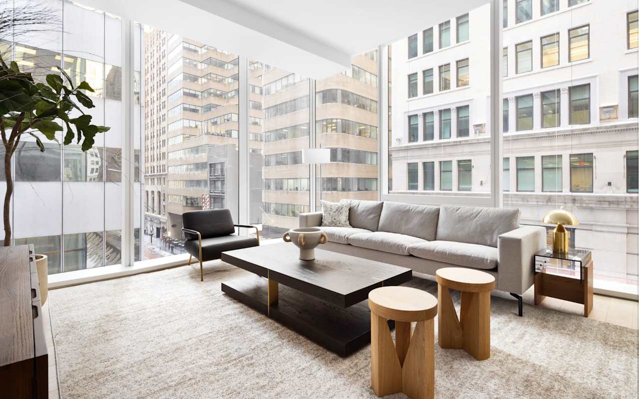 A modern living room with large windows, a gray sofa, black chair, wooden coffee table, and city views.